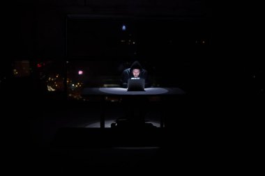 Genç yetenekli hacker karanlık ofiste çalışırken dizüstü bilgisayar kullanıyor. Arka planda büyük şehir ışıkları var.