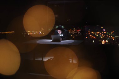 Genç yetenekli hacker karanlık ofiste çalışırken dizüstü bilgisayar kullanıyor. Arka planda büyük şehir ışıkları var.