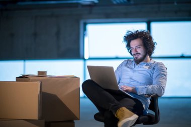 İnşaat alanındaki genç erkek mimar yeni açılan ofiste yanında karton kutularla dizüstü bilgisayar kullanarak belge ve iş akışını kontrol ediyor.