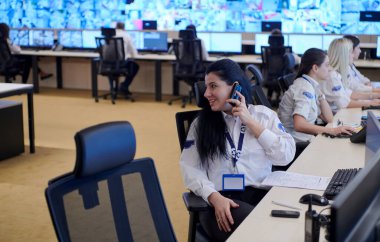 Kadın güvenlik görevlisi birden fazla monitörde çalışan güvenlik görevlileriyle çalışırken telefonda konuşuyor.