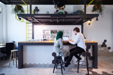 Uluslararası çok kültürlü iş adamları. Elma yiyen Afrika 'lı Müslüman kadın yeşil başörtüsü takıyor, çay içiyor ve modern açık planlama ofisinin rahatlama bölümünde dizüstü bilgisayarda birlikte çalışıyoruz.