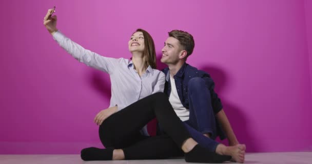 Ritratto di coppia caucasica positiva in abiti colorati divertendosi mentre scattava foto di selfie sul cellulare su sfondo rosa — Video Stock