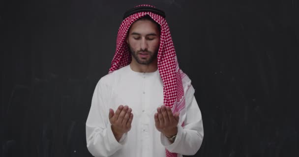 伝統的な服を着た若いアラビア人男性が伝統的な神への祈りをし 現代のイスラム教のファッションとラマダーン カレームの概念を表す黒い黒板の前で手をつないで祈り続ける — ストック動画