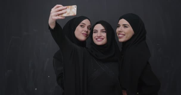 現代のイスラムファッション技術とラマダーン カレムの概念を代表する黒い黒板の前で自撮りをしながら携帯電話を使ってヒジャーブを着た若い美しいイスラム教徒の女性のグループ — ストック動画