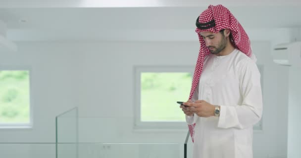 家庭でスマートフォンを使いながら伝統的な衣服を身に着けている若い現代のイスラム教徒のアラビア人ビジネスマン — ストック動画