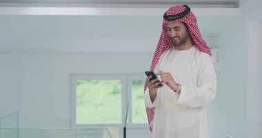 Genç ve modern Müslüman Arap iş adamı geleneksel kıyafetler giyerken akıllı telefon kullanıyor.