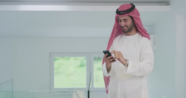 家庭でスマートフォンを使いながら伝統的な衣服を身に着けている若い現代のイスラム教徒のアラビア人ビジネスマン — ストック動画