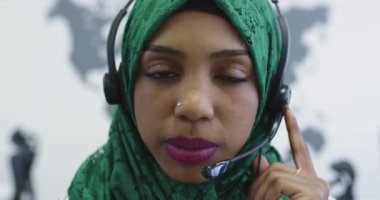 Yeşil başörtülü Afrikalı Müslüman kadın müşteri temsilcisi telefon kulaklıklı iş kadını modern çağrı merkezindeki müşterilere internet üzerinden yardım ve destek veriyor.