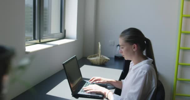 Jovem empresária alegre atraente trabalhando no laptop e sorrindo enquanto se senta em sua mesa em um escritório moderno brilhante — Vídeo de Stock