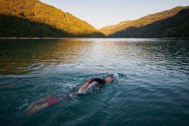 Triatlon sporcusu gün doğumunda gölde yüzüyor. Dalgıç kıyafeti giyiyor.