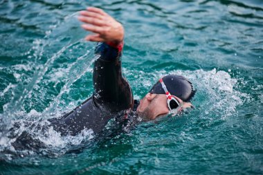 Triatlon sporcusu gün doğumunda gölde yüzüyor. Dalgıç kıyafeti giyiyor.