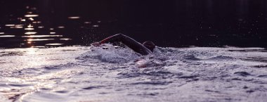 Triatlon sporcusu gün doğumunda gölde yüzüyor. Dalgıç kıyafeti giyiyor.