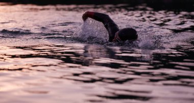 Triatlon sporcusu gün doğumunda gölde yüzüyor. Dalgıç kıyafeti giyiyor.