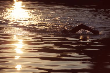 Triatlon sporcusu gün doğumunda gölde yüzüyor. Dalgıç kıyafeti giyiyor.