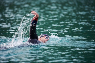 Triatlon sporcusu gün doğumunda gölde yüzüyor. Dalgıç kıyafeti giyiyor.