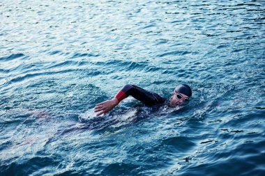 Triatlon sporcusu gün doğumunda gölde yüzüyor. Dalgıç kıyafeti giyiyor.