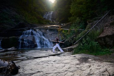 Yoga yapan kadın, gün doğumunda şelalenin yanında poz veriyor. Açık havada sağlıklı meditasyon yapıyor. Rahatlama farkındalık ahenk ruhsal geri çekilme sükunet konsepti.