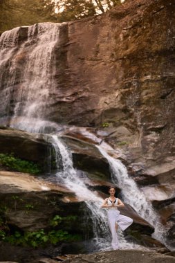 Gün doğumunda şelalenin önünde yoga ağacı pozu veren bir kadın doğada huzurlu bir meditasyon yapıyor. Sakin ve rahatlatıcı ağır çekim sahnesi