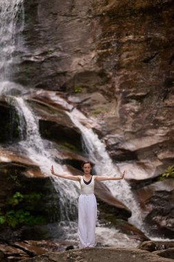Güzel genç bir kadın gün batımında şelale ve nehir kenarında ağır ağır tai chi hareketleri yapıyor. Doğadaki uyum dengesini ve uygunluğu yakalıyor.