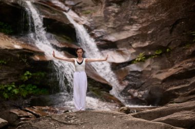 Güzel genç bir kadın gün batımında şelale ve nehir kenarında ağır ağır tai chi hareketleri yapıyor. Doğadaki uyum dengesini ve uygunluğu yakalıyor.