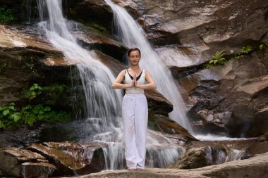 Güzel genç bir kadın gün batımında şelale ve nehir kenarında ağır ağır tai chi hareketleri yapıyor. Doğadaki uyum dengesini ve uygunluğu yakalıyor.