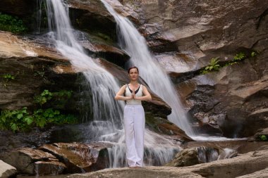 Güzel genç bir kadın gün batımında şelale ve nehir kenarında ağır ağır tai chi hareketleri yapıyor. Doğadaki uyum dengesini ve uygunluğu yakalıyor.