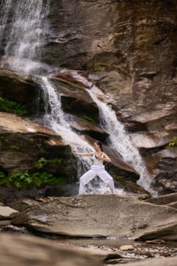 Güzel genç bir kadın gün batımında şelale ve nehir kenarında ağır ağır tai chi hareketleri yapıyor. Doğadaki uyum dengesini ve uygunluğu yakalıyor.