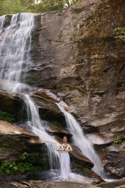Güzel genç bir kadın gün batımında şelale ve nehir kenarında ağır ağır tai chi hareketleri yapıyor. Doğadaki uyum dengesini ve uygunluğu yakalıyor.