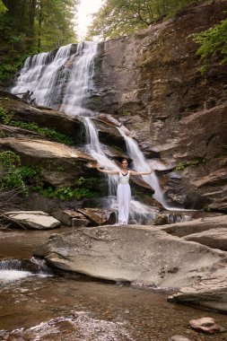 Güzel genç bir kadın gün batımında şelale ve nehir kenarında ağır ağır tai chi hareketleri yapıyor. Doğadaki uyum dengesini ve uygunluğu yakalıyor.