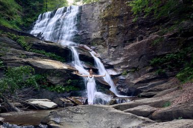 Güzel genç bir kadın gün batımında şelale ve nehir kenarında ağır ağır tai chi hareketleri yapıyor. Doğadaki uyum dengesini ve uygunluğu yakalıyor.