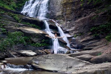 Güzel genç bir kadın gün batımında şelale ve nehir kenarında ağır ağır tai chi hareketleri yapıyor. Doğadaki uyum dengesini ve uygunluğu yakalıyor.