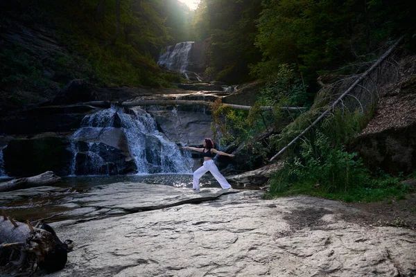 Yoga yapan kadın, gün doğumunda şelalenin yanında poz veriyor. Açık havada sağlıklı meditasyon yapıyor. Rahatlama farkındalık ahenk ruhsal geri çekilme sükunet konsepti.