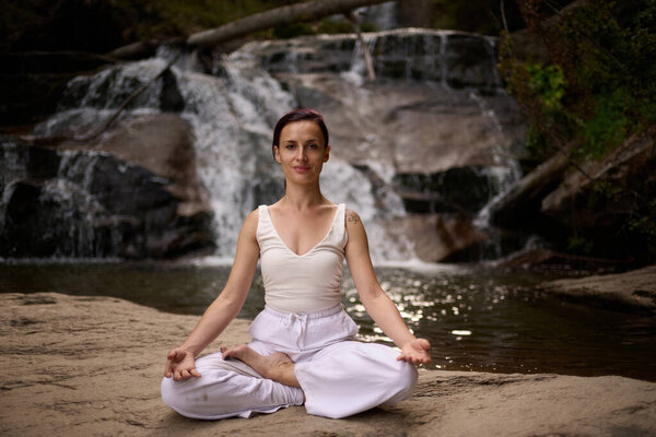 Young woman sitting in lotus pose under tropical waterfall meditating with calm strength and mindfulness fully connected to nature energy tranquility and inner balance