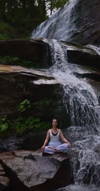 Lotus 'ta oturan kadın, tropikal şelale altında poz veriyor sakin bir güç ve farkındalık ile doğa enerjisi sükuneti ve iç dengesi ile bütünüyle bağlantılı. Ağır çekimin dikey videosu
