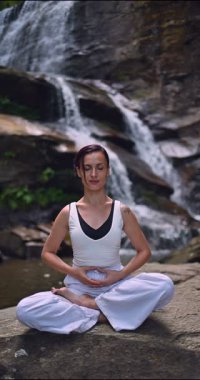 Lotus 'ta oturan kadın, tropikal şelale altında poz veriyor sakin bir güç ve farkındalık ile doğa enerjisi sükuneti ve iç dengesi ile bütünüyle bağlantılı. Ağır çekimin dikey videosu