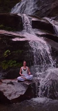 Lotus 'ta oturan kadın, tropikal şelale altında poz veriyor sakin bir güç ve farkındalık ile doğa enerjisi sükuneti ve iç dengesi ile bütünüyle bağlantılı. Ağır çekimin dikey videosu