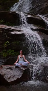 Lotus 'ta oturan kadın, tropikal şelale altında poz veriyor sakin bir güç ve farkındalık ile doğa enerjisi sükuneti ve iç dengesi ile bütünüyle bağlantılı. Ağır çekimin dikey videosu