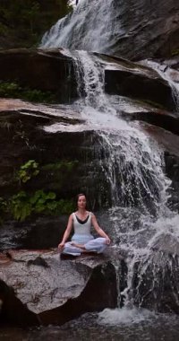 Lotus 'ta oturan kadın, tropikal şelale altında poz veriyor sakin bir güç ve farkındalık ile doğa enerjisi sükuneti ve iç dengesi ile bütünüyle bağlantılı. Ağır çekimin dikey videosu
