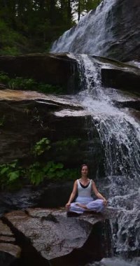Lotus 'ta oturan kadın, tropikal şelale altında poz veriyor sakin bir güç ve farkındalık ile doğa enerjisi sükuneti ve iç dengesi ile bütünüyle bağlantılı. Ağır çekimin dikey videosu