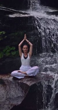 Lotus 'ta oturan kadın, tropikal şelale altında poz veriyor sakin bir güç ve farkındalık ile doğa enerjisi sükuneti ve iç dengesi ile bütünüyle bağlantılı. Ağır çekimin dikey videosu