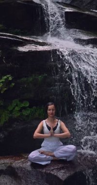 Lotus 'ta oturan kadın, tropikal şelale altında poz veriyor sakin bir güç ve farkındalık ile doğa enerjisi sükuneti ve iç dengesi ile bütünüyle bağlantılı. Ağır çekimin dikey videosu