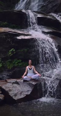 Lotus 'ta oturan kadın, tropikal şelale altında poz veriyor sakin bir güç ve farkındalık ile doğa enerjisi sükuneti ve iç dengesi ile bütünüyle bağlantılı. Ağır çekimin dikey videosu