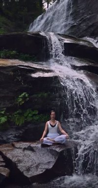 Lotus 'ta oturan kadın, tropikal şelale altında poz veriyor sakin bir güç ve farkındalık ile doğa enerjisi sükuneti ve iç dengesi ile bütünüyle bağlantılı. Ağır çekimin dikey videosu