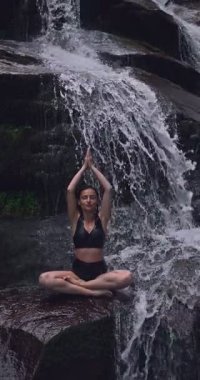 Lotus 'ta oturan kadın, tropikal şelale altında poz veriyor sakin bir güç ve farkındalık ile doğa enerjisi sükuneti ve iç dengesi ile bütünüyle bağlantılı. Ağır çekimin dikey videosu