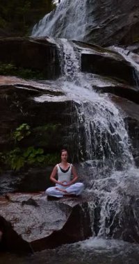 Lotus 'ta oturan kadın, tropikal şelale altında poz veriyor sakin bir güç ve farkındalık ile doğa enerjisi sükuneti ve iç dengesi ile bütünüyle bağlantılı. Ağır çekimin dikey videosu