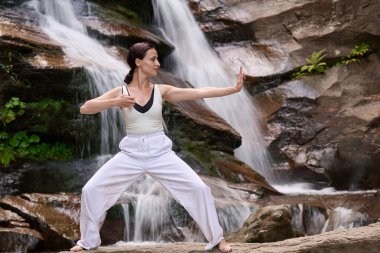 Güzel genç bir kadın gün batımında şelale ve nehir kenarında ağır ağır tai chi hareketleri yapıyor. Doğadaki uyum dengesini ve uygunluğu yakalıyor.