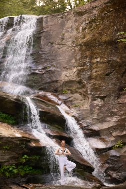 Gün doğumunda şelalenin önünde yoga ağacı pozu veren bir kadın doğada huzurlu bir meditasyon yapıyor. Sakin ve rahatlatıcı ağır çekim sahnesi