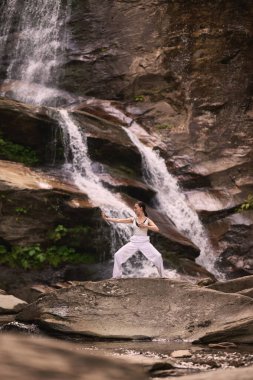 Güzel genç bir kadın gün batımında şelale ve nehir kenarında ağır ağır tai chi hareketleri yapıyor. Doğadaki uyum dengesini ve uygunluğu yakalıyor.
