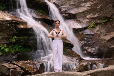 Güzel genç bir kadın gün batımında şelale ve nehir kenarında ağır ağır tai chi hareketleri yapıyor. Doğadaki uyum dengesini ve uygunluğu yakalıyor.