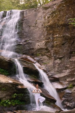Gün doğumunda şelalenin önünde yoga ağacı pozu veren bir kadın doğada huzurlu bir meditasyon yapıyor. Sakin ve rahatlatıcı ağır çekim sahnesi